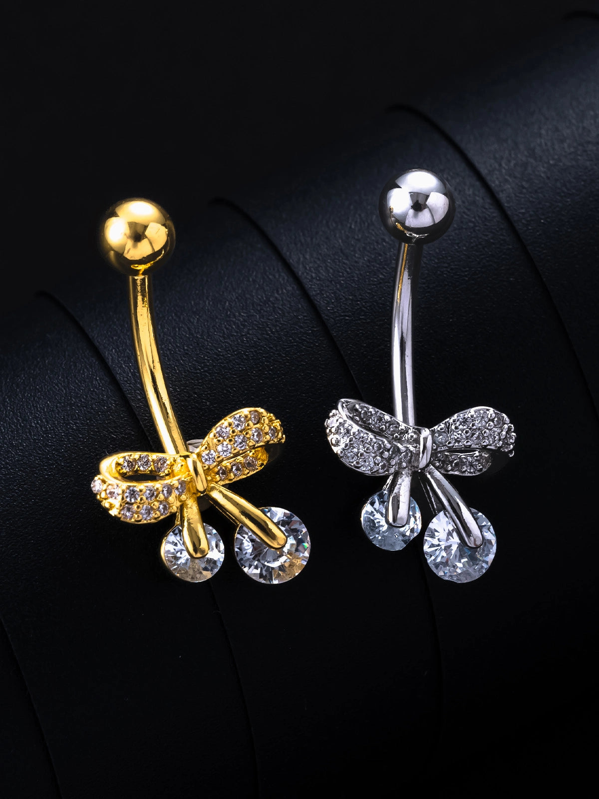A set of belly ring product photos in gold and silver. Each piece features an integrated bow design at the base, fully set with small diamonds.
